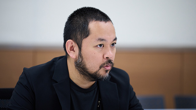 Tatsuya Ishikawa, wearing a black jacket, giving an interview.