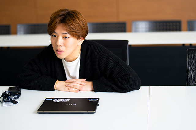 机に手を置いて話すHOMARE