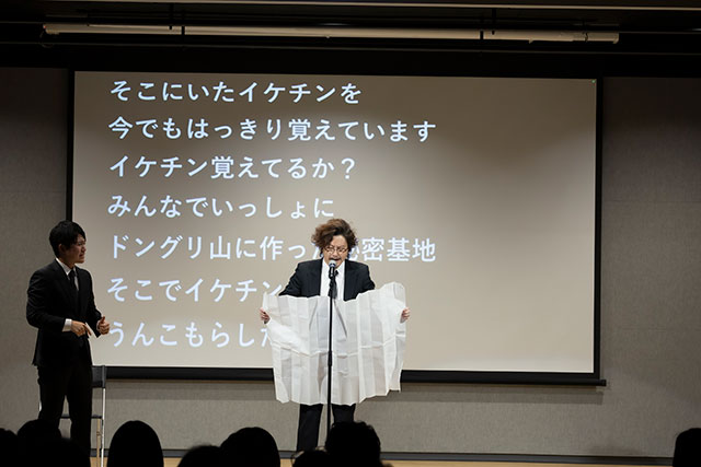 よれよれの喪服を着て、大きな紙を広げて、手紙を読むマツモトクラブ